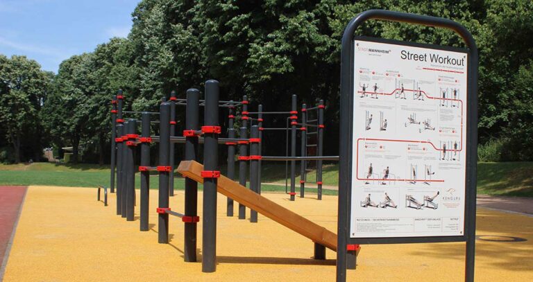 Calisthenics-Anlage im Unteren Luisenpark Mannheim mit gelbem Fallschutzboden, schwarzen und roten Trainingsgeräten sowie Infotafel mit Übungsanleitungen von Kenguru Pro vor grüner Parkkulisse.