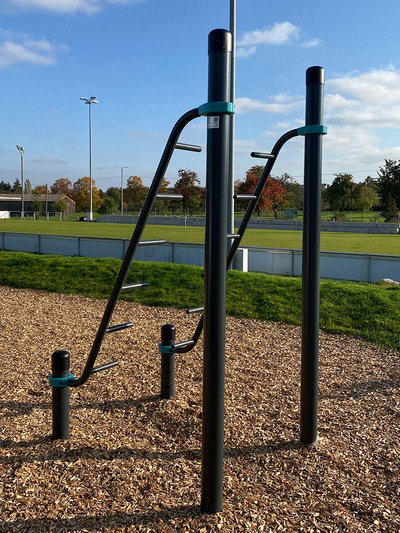 Outdoor-Trainingsgerät mit gebogenen Griffstangen für Liegestütze und Armzüge, installiert auf Holzhackschnitzel-Untergrund mit Sportplatz im Hintergrund