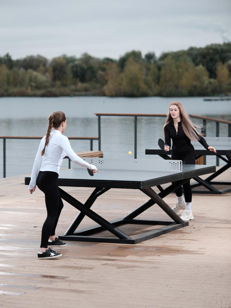 Zwei junge Frauen spielen Tischtennis an einer modernen Outdoor-Tischtennisplatte von Kenguru Pro mit Seeblick