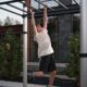 Ein Jugendlicher trainiert an einer Hangel­leiter einer modernen Outdoor-Calisthenics-Anlage. Mit ausgestreckten Armen greift er fest an die Sprossen, um sich kraftvoll voranzuhangeln. Die Anlage aus Metall ist für funktionelles Training im Freien konzipiert und steht vor einem modernen Gebäude, umgeben von grünen Pflanzen. Ideal für Koordination, Griffkraft und Ganzkörpertraining.