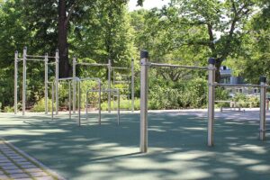Calisthenics Park Berlin Zeppelinplatz mit Edelstahl-Trainingsgeräten auf einem grünen Gummiboden. Zu sehen sind verschiedene Trainingsstationen wie Klimmzugstangen, Barren und eine Sprossenwand. Die Anlage befindet sich im Freien, umgeben von Bäumen und Grünflächen, mit einem Gehweg aus Pflastersteinen im Vordergrund.