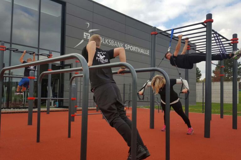 Mehrere Personen trainieren an einem Calisthenics Park Deißlingen mit dunkelgrauen Metallstangen und roten Verbindungsstellen vor der Volksbank Sporthalle. Die Sportler nutzen verschiedene Geräte wie Barren, Monkey Bars und Sling-Trainer für Übungen wie Dips, Klimmzüge und TRX-Training. Der Boden ist mit einem roten Fallschutzbelag versehen.