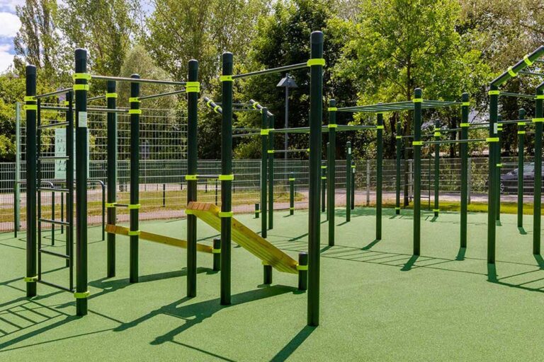 Calisthenics Park Königs Wusterhausen mit mehreren dunkelgrünen Metallstangen und gelben Verbindungsringen auf einem grünen Gummiboden. Zu sehen sind verschiedene Trainingsstationen wie Klimmzugstangen, Barren, Sprossenwände und eine schräg montierte Holzbank.