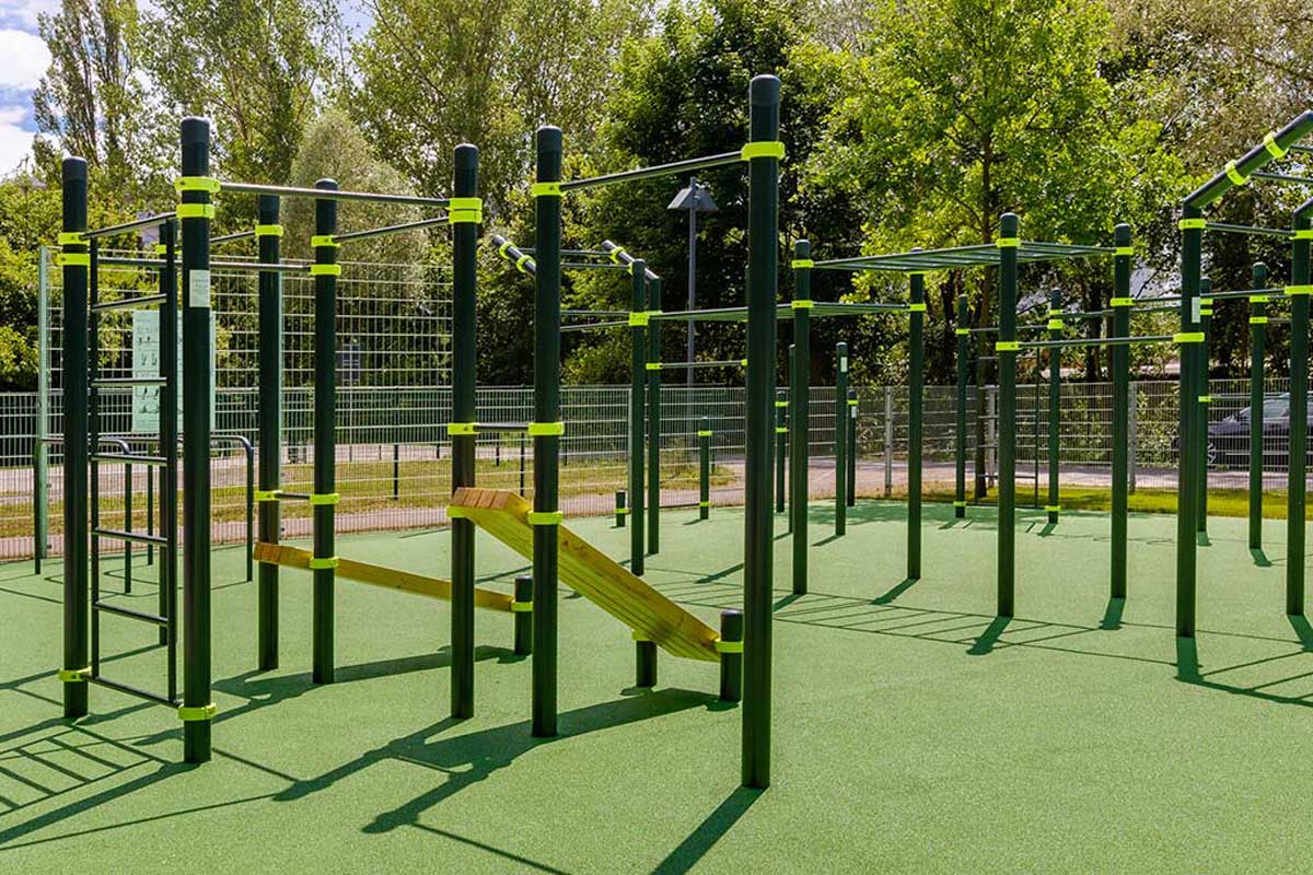 Calisthenics Park Königs Wusterhausen mit mehreren dunkelgrünen Metallstangen und gelben Verbindungsringen auf einem grünen Gummiboden. Zu sehen sind verschiedene Trainingsstationen wie Klimmzugstangen, Barren, Sprossenwände und eine schräg montierte Holzbank.