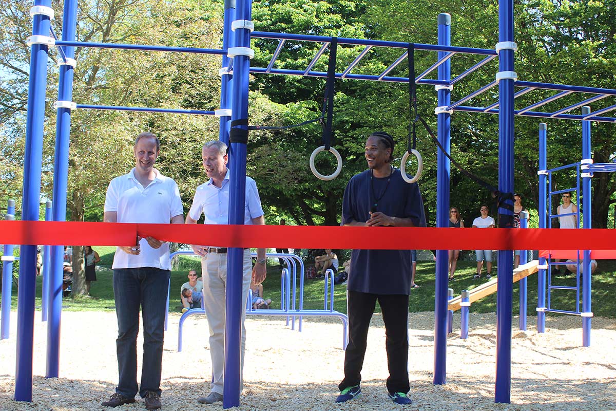 Eröffnung eines Calisthenics Park Ravensburg mit blauen Trainingsgeräten und Holzhackschnitzelboden. Drei Männer stehen vor einem roten Band, das symbolisch durchgeschnitten wird. Im Hintergrund sind verschiedene Sportgeräte wie Monkey Bars und Ringe sowie Zuschauer zu sehen. Die Veranstaltung findet bei sonnigem Wetter im Grünen statt.