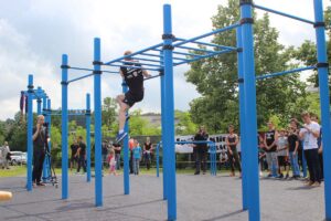 Sportveranstaltung in einem Calisthenics Workout Park Rimbach mit blauen Trainingsgeräten. Im Vordergrund führt ein junger Mann eine Übung an einer Monkey-Bar-Anlage aus, während Zuschauer, darunter Kinder und Erwachsene, die Aktion beobachten. Einige Personen filmen oder fotografieren das Geschehen. Im Hintergrund sind weitere Geräte, ein Kameramann und eine grüne Parklandschaft zu sehen.