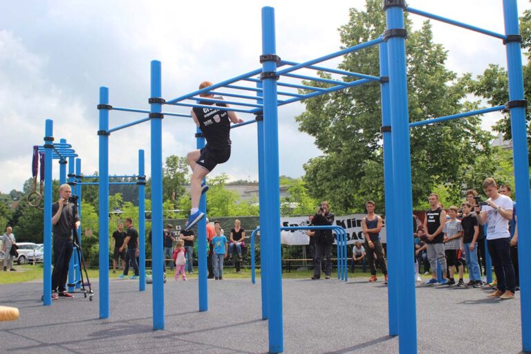 Sportveranstaltung in einem Calisthenics Workout Park Rimbach mit blauen Trainingsgeräten. Im Vordergrund führt ein junger Mann eine Übung an einer Monkey-Bar-Anlage aus, während Zuschauer, darunter Kinder und Erwachsene, die Aktion beobachten. Einige Personen filmen oder fotografieren das Geschehen. Im Hintergrund sind weitere Geräte, ein Kameramann und eine grüne Parklandschaft zu sehen.