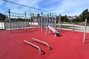 Calisthenics Park von KENGURU PRO mit mehreren Klimmzugstangen, Sprossenwand, Turnringen, Schrägbank auf rotem Fallschutzboden, in einer herbstlichen Sportanlage.
