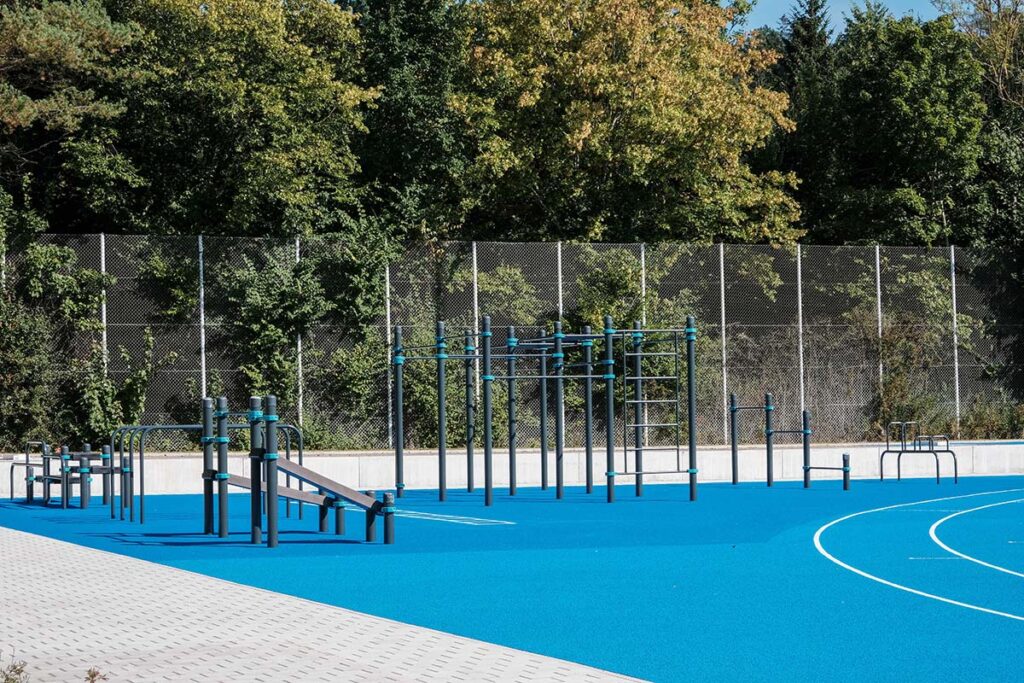 Moderner Calisthenics Park in Villingen-Schwenningen mit blauen Fallschutzboden, Klimmzugstangen, Barren und Hangelleiter vor bewaldeter Kulisse.
