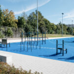 Der moderne Calisthenics Park in Villingen-Schwenningen mit blauen Gummiböden, Klimmzugstangen, Barren und einer angrenzenden Laufbahn vor einer dichten Waldkulisse unter blauem Himmel