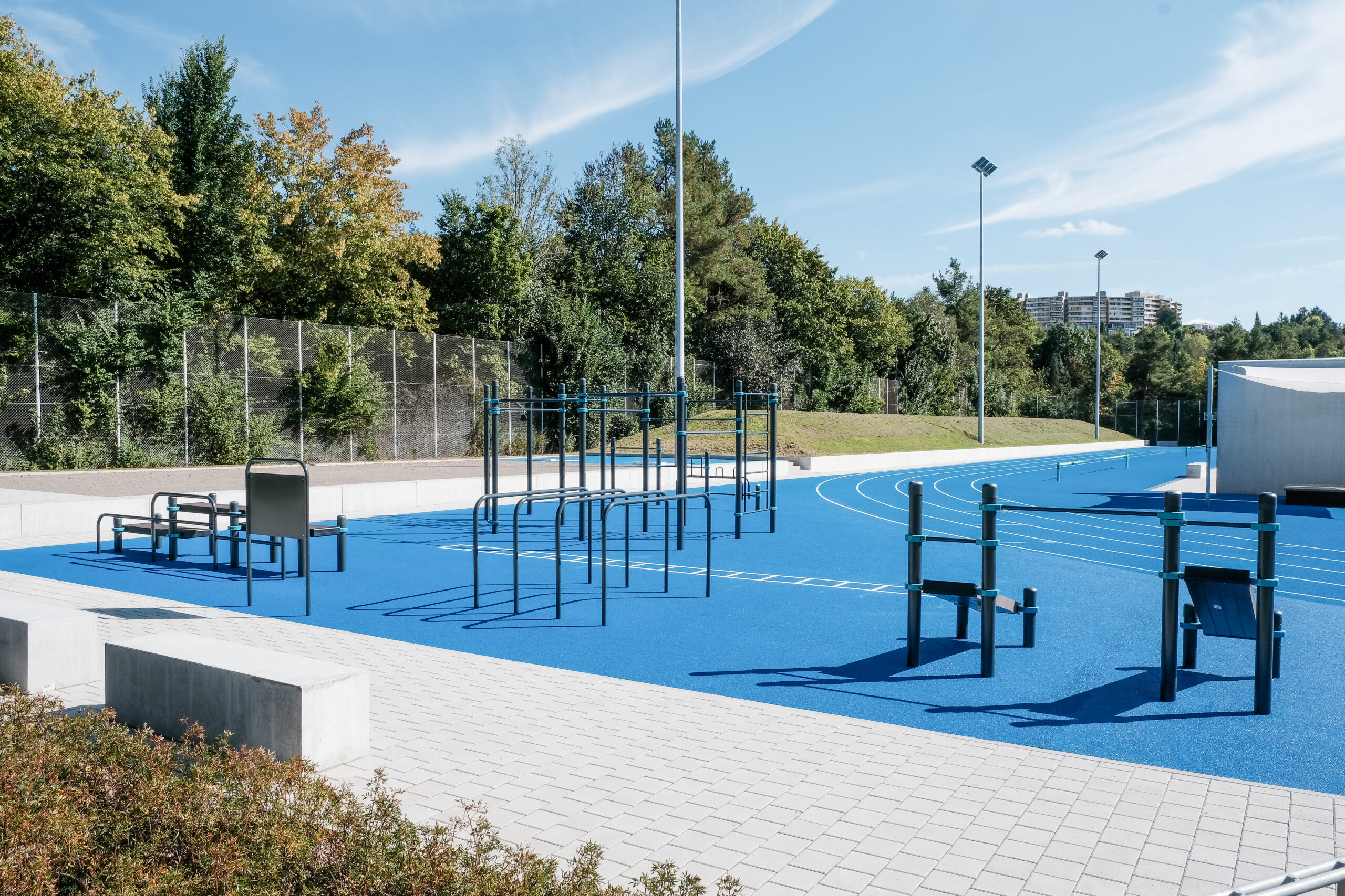 Der moderne Calisthenics Park in Villingen-Schwenningen mit blauen Gummiböden, Klimmzugstangen, Barren und einer angrenzenden Laufbahn vor einer dichten Waldkulisse unter blauem Himmel