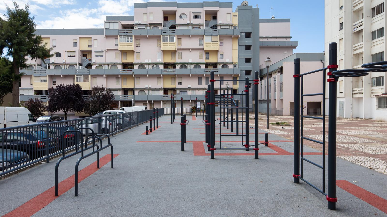 Calisthenics Anlage von KENGURU PRO zwischen Wohnblöcken in Lissabon mit Reckstangen, Sprossenwand und Barren auf grau-rotem Fallschutzbelag