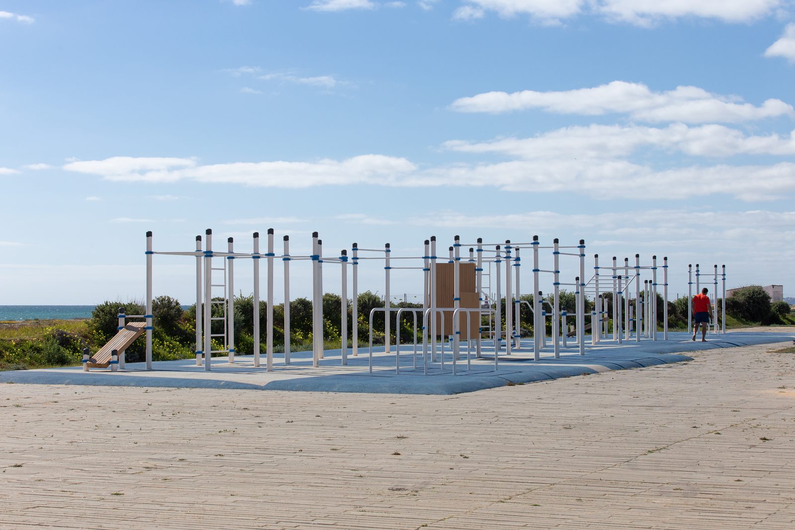 Bewegungsparcours von KENGURU PRO entlang einer Strandpromenade: lange Reihe von Stationen mit Reckstangen, Barren, Sprossenwand und Schrägbank auf blauem Belag am Meer