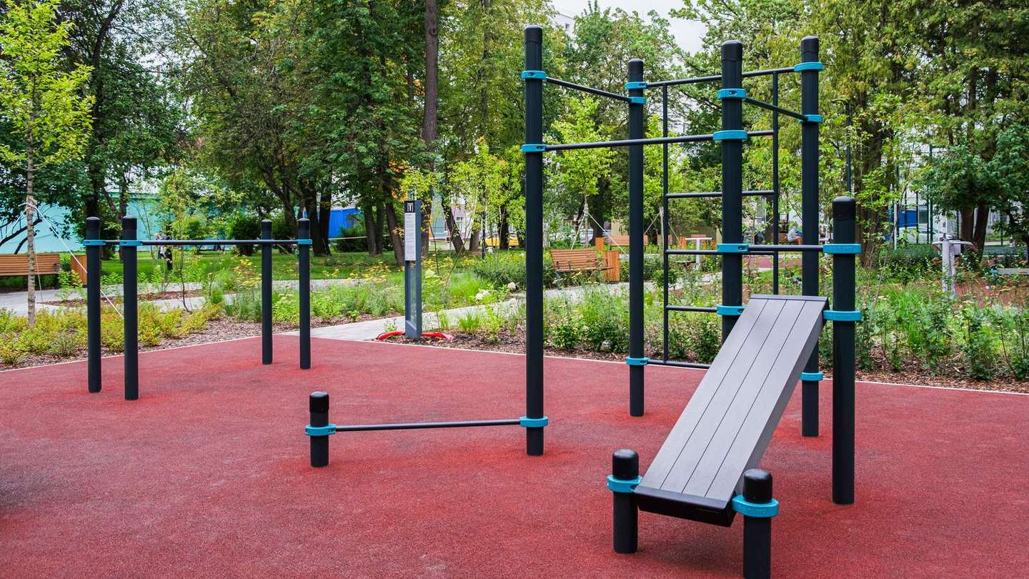 Outdoor Gym von KENGURU PRO mit Trainingsbänken, Sprossenwand und Reckstangen auf rotem Fallschutzbelag im Grünen