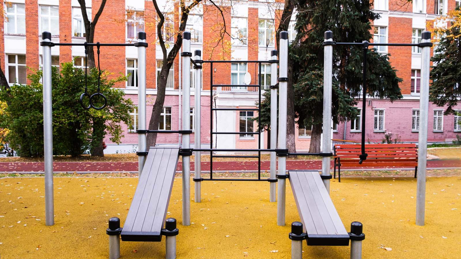 Bewegungspark im Herbst mit Calisthenics Station von KENGURU PRO: zwei Schrägbänke, Turnringe und Sprossenwand auf gelbem Belag, daneben Parkbank und Bäume